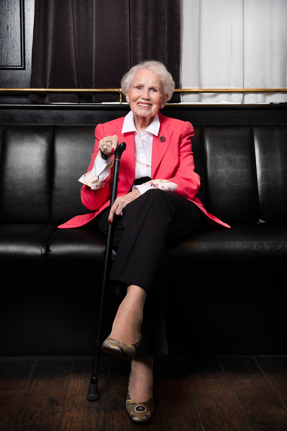 Marguerite, the grandmother behind Atelier Marguerite, smiling on a black leather sofa in a pink jacket
